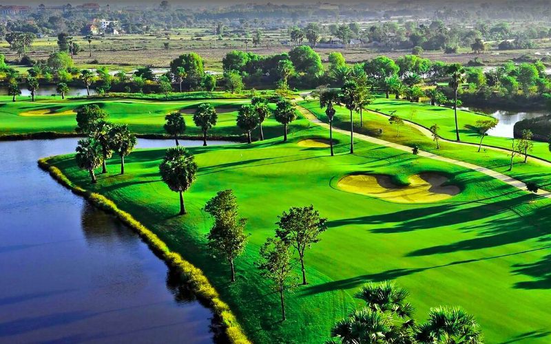 Vue aérienne d’un parcours de golf luxuriant avec des fairways bordés de palmiers, de bunkers de sable doré et de plans d’eau sinueux, dans un paysage tropical baigné de lumière matinale