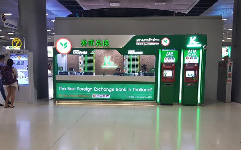 Bureau de change de la Kasikornbank à l’aéroport en Thaïlande, avec deux distributeurs automatiques verts pour retirer des bahts thaïlandais et un comptoir de change affichant les taux de devises.