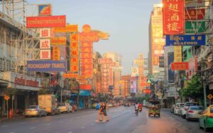 Rue animée du quartier de Chinatown à Bangkok en fin d’après-midi, avec des enseignes colorées en thaï et en chinois, quelques passants traversant la rue et des véhicules garés le long des trottoirs