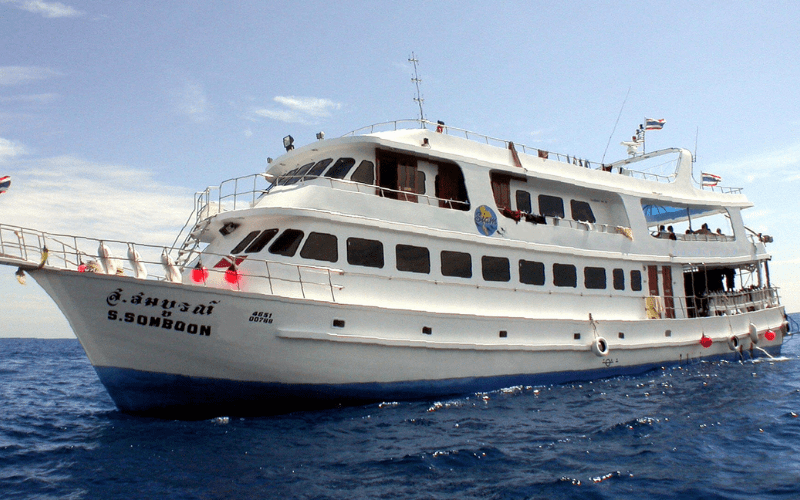 Le bateau blanc navigue sur une mer calme sous un ciel bleu. Le bateau a plusieurs ponts et des fenêtres alignées sur ses côtés. Des drapeaux thaïlandais flottent sur le bateau, et il y a des bouées rouges accrochées sur les côtés.