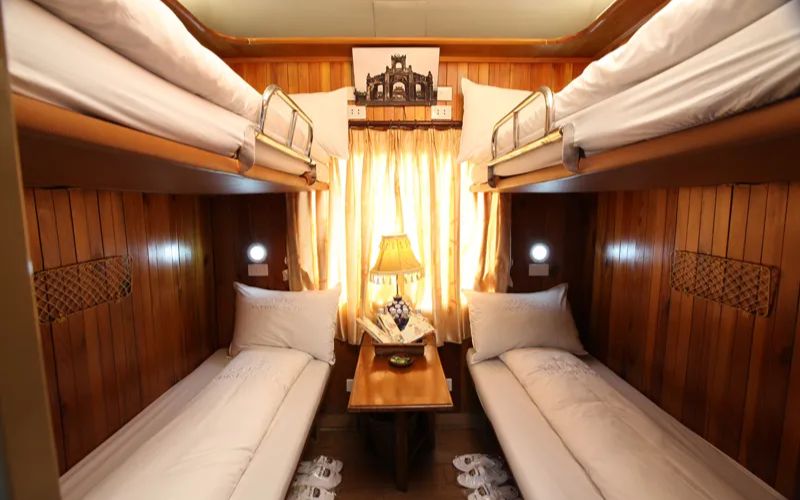 Cabine en bois chaleureuse d’un train de nuit au Vietnam, équipée de quatre couchettes confortables superposées, d’une petite table avec lampe décorative et d’une fenêtre aux rideaux dorés laissant entrer la lumière