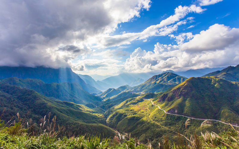 Vue panoramique sur les montagnes verdoyantes de Sapa, avec une route sinueuse serpentant à travers les vallées et des rayons de soleil perçant les nuages, illuminant le paysage majestueux