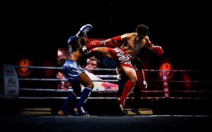 Combat de Muay Thai au stade Rajadamnern, montrant un moment intense où un combattant en rouge exécute un coup de pied spectaculaire contre son adversaire en bleu