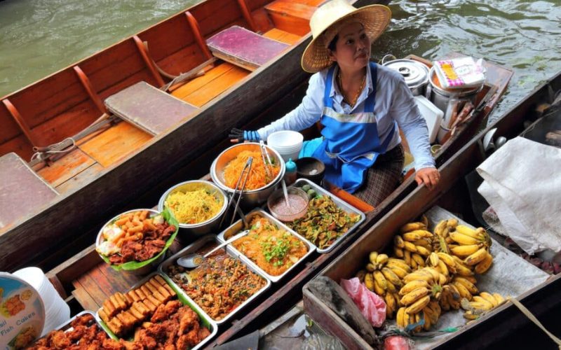 Une vendeuse sur une barque traditionnelle au marché flottant, proposant des plats thaïlandais colorés, des fruits frais et des spécialités locales