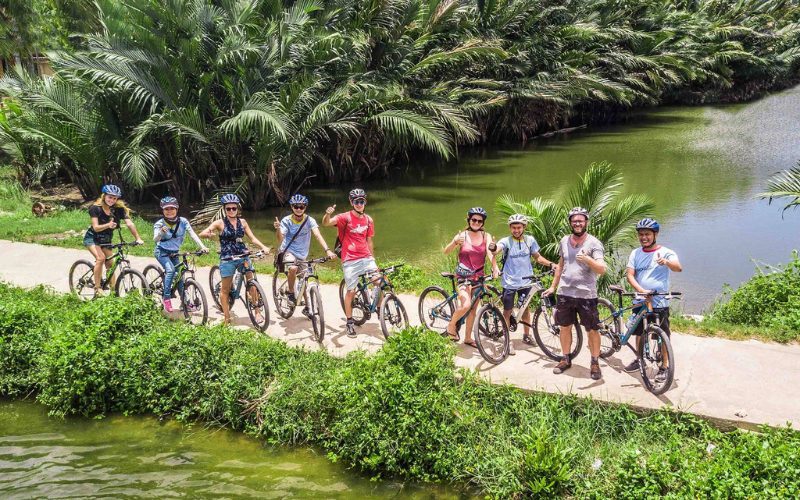 Groupe de cyclistes explorant la campagne de Hoi An, pédalant le long d’un sentier bordé de palmiers et de canaux verdoyants