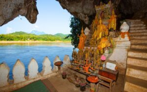 Intérieur des grottes de Pak Ou à Luang Prabang, avec des statues de Bouddha dorées ornées de guirlandes, surplombant le fleuve Mékong et les montagnes verdoyantes en arrière-plan