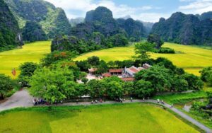 Vue d’ensemble d’un paysage rural de Ninh Binh, avec des rizières lumineuses, des montagnes karstiques recouvertes de végétation et un petit ensemble de bâtiments traditionnels niché au cœur de la campagne