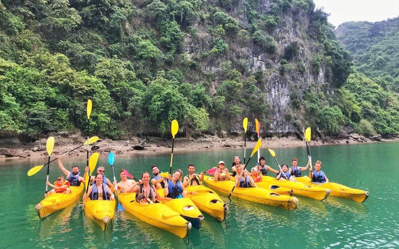 Groupe de touristes souriants en kayak jaune sur les eaux vert émeraude de Cat Ba, entourés de falaises couvertes de végétation luxuriante