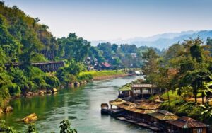 Vue panoramique sur la rivière Kwai bordée de maisons flottantes et entourée de collines verdoyantes, avec une voie ferrée historique visible à flanc de montagne