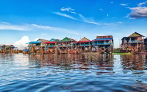 Maisons colorées sur pilotis bordant le lac Tonlé Sap au Cambodge, se reflétant dans l’eau calme sous un ciel bleu parsemé de nuages, typique des villages flottants de la région