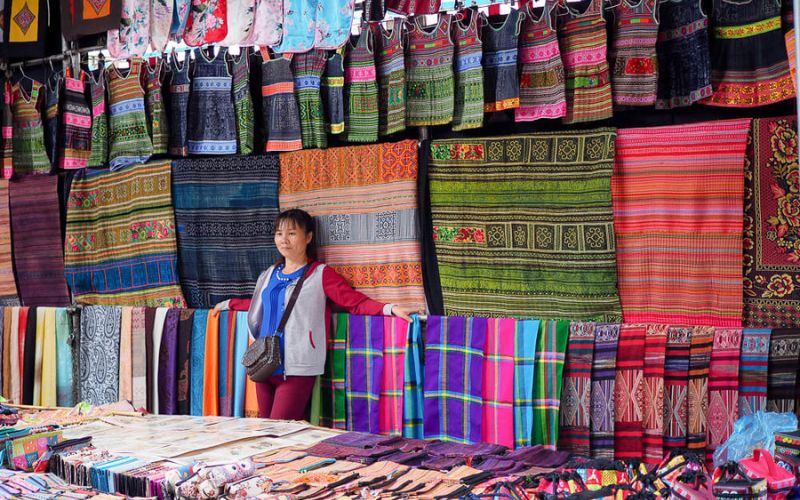 Marchande au marché de Sapa devant son étal coloré de tissus et d’étoffes traditionnelles des ethnies locales, aux motifs variés et aux couleurs vives
