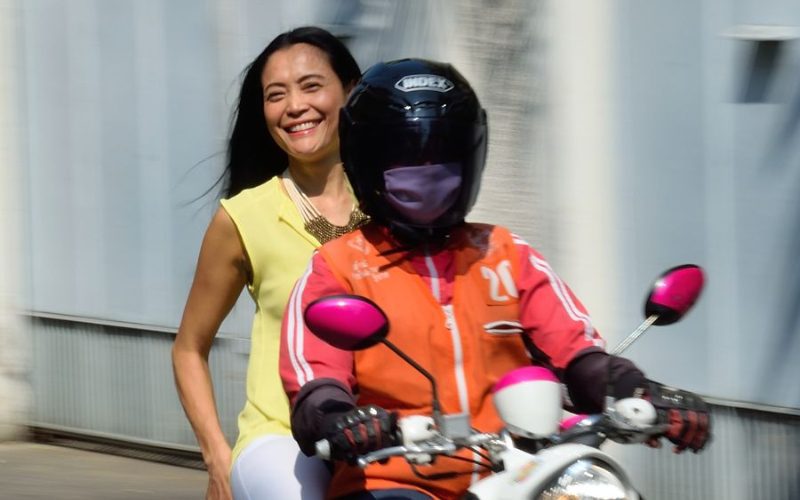 Femme souriante en tenue jaune assise à l'arrière d'un taxi moto à Bangkok, conduit par un chauffeur portant un casque noir et une veste orange