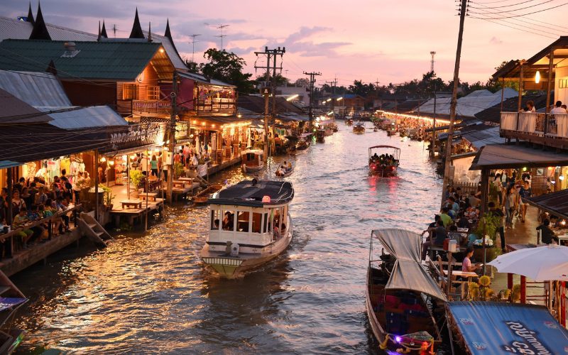 Des bateaux naviguent sur le canal, tandis que les rives sont bordées de maisons traditionnelles, de restaurants et d'étals éclairés, créant une ambiance chaleureuse et vibrante