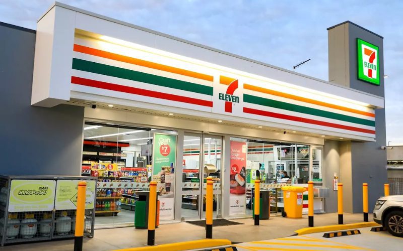 Façade d’un magasin 7-Eleven moderne et bien éclairé en soirée, avec des vitrines transparentes montrant les produits à l’intérieur et un parking aménagé à l’avant