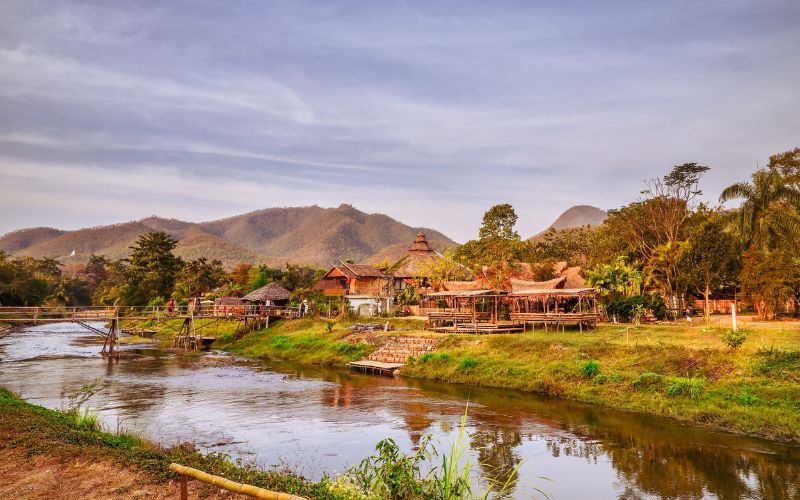 Village traditionnel du nord de la Thaïlande au bord d’une rivière, entouré de montagnes verdoyantes et de maisons en bois