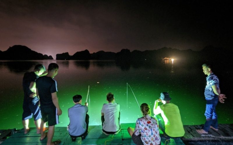 Groupe de personnes pêchant le calmar de nuit sur un quai éclairé, avec vue sur les montagnes sombres et la mer calme de Cat Ba