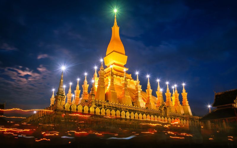 Vue nocturne spectaculaire du stupa doré de Pha That Luang à Vientiane, Laos, illuminé de lumières vives avec un ciel sombre en arrière-plan et des traînées lumineuses de fidèles en procession au premier plan
