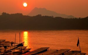 Coucher de soleil sur le fleuve Mékong à Luang Prabang, avec des bateaux traditionnels en bois et des montagnes en arrière-plan