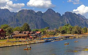 Paysage pittoresque de Vang Vieng, au Laos, avec des maisons traditionnelles en bord de rivière, des montagnes karstiques en arrière-plan et des touristes faisant du kayak sur la rivière Nam Song