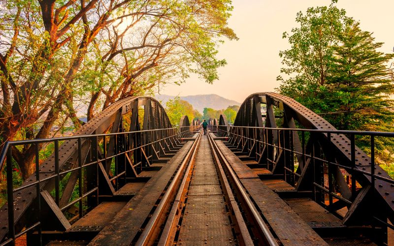 Pont en acier de la rivière Kwai à Kanchanaburi, baigné par la lumière dorée du coucher de soleil, avec des visiteurs marchant sur les rails historiques