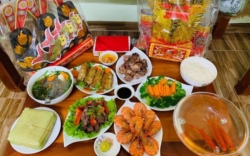 Table d’offrandes pour les Génies du Foyer (Ông Công, Ông Táo) au Vietnam, comprenant des plats traditionnels comme le bánh chưng, les crevettes, les rouleaux frits, les légumes, le riz et un bol contenant des carpes vivantes destinées au rituel