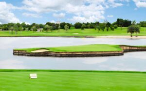 Terrain de golf Royal Gems Golf City - Dream Arena avec un green insulaire entouré d'eau, un paysage verdoyant et un ciel dégagé en arrière-plan