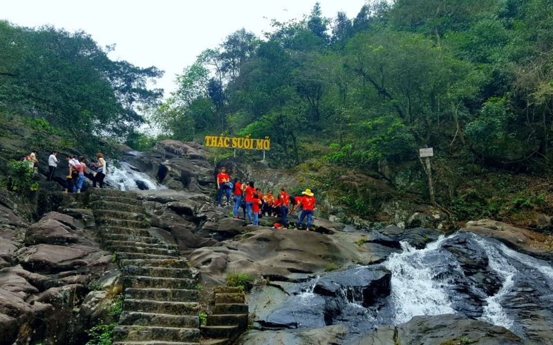 Groupe de visiteurs vêtus de t-shirts rouges ornés d’étoiles jaunes explorant la cascade de Suối Mỡ, entourée de rochers et de végétation dense