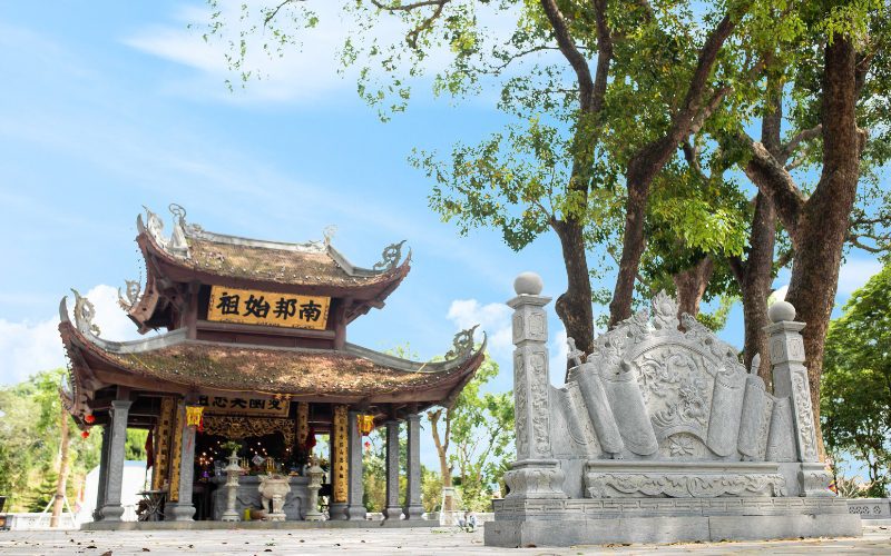Temple et tombeau du roi Kinh Dương Vương à Bắc Ninh, avec un bâtiment en bois traditionnel au toit recourbé couvert de mousse, entouré d’arbres anciens et d’ornements en pierre finement sculptés