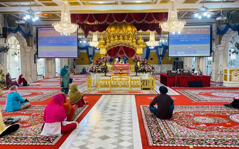 Intérieur du temple sikh Gurudwara Sri Guru Singh Sabha à Bangkok, avec des fidèles assis sur des tapis colorés, face à l’autel doré richement décoré