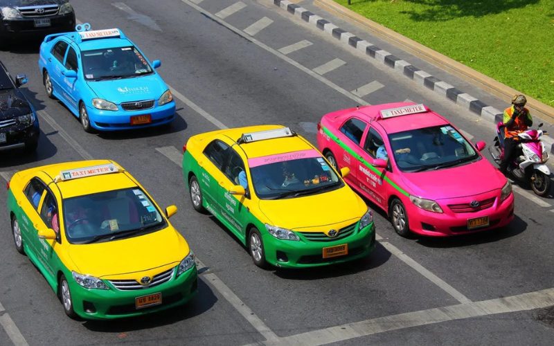 Taxis colorés de Bangkok, incluant des véhicules jaunes et verts, roses et bleus, circulant sur une route animée, avec un motard en taxi moto à proximité