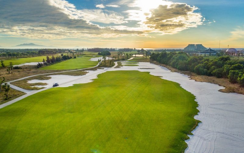 Vue aérienne d’un parcours de golf verdoyant au lever du soleil, avec bunkers de sable blanc, allées sinueuses et bâtiments modernes en arrière-plan
