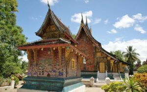 Vue du temple Wat Xieng Thong à Luang Prabang, au Laos, avec ses toits traditionnels superposés, ses façades richement décorées de mosaïques colorées et d’or, entouré de végétation tropicale sous un ciel bleu éclatant