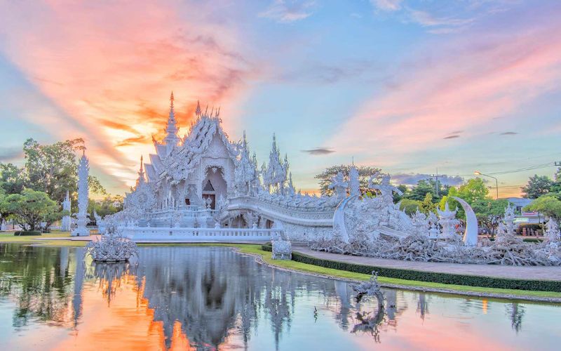 Vue spectaculaire du Temple Blanc (Wat Rong Khun) à Chiang Rai, reflété dans un étang tranquille sous un ciel aux teintes pastel