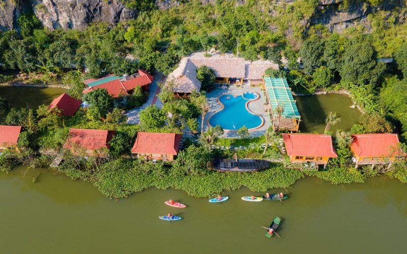 Vue aérienne d’un écolodge au bord d’une rivière à Phu Tho, avec bungalows aux toits rouges, piscine entourée de verdure et visiteurs pratiquant le kayak et le paddle