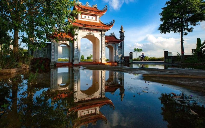 Porte d’entrée traditionnelle du site historique de Lê Chi Viên à Bắc Ninh, avec son reflet dans une flaque d’eau, entourée d’arbres verdoyants sous un ciel bleu lumineux